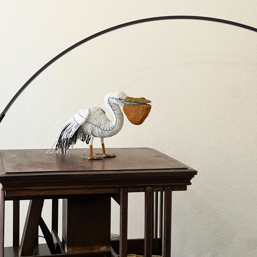 Pelican figurine in white knit with orange beak and tassel feet perched on a wooden shelf.