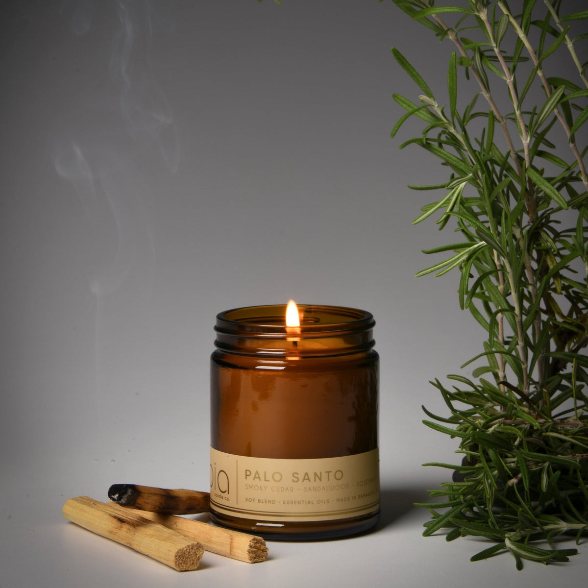 Amber candle jar with lit flame and palo wood pieces near greenery