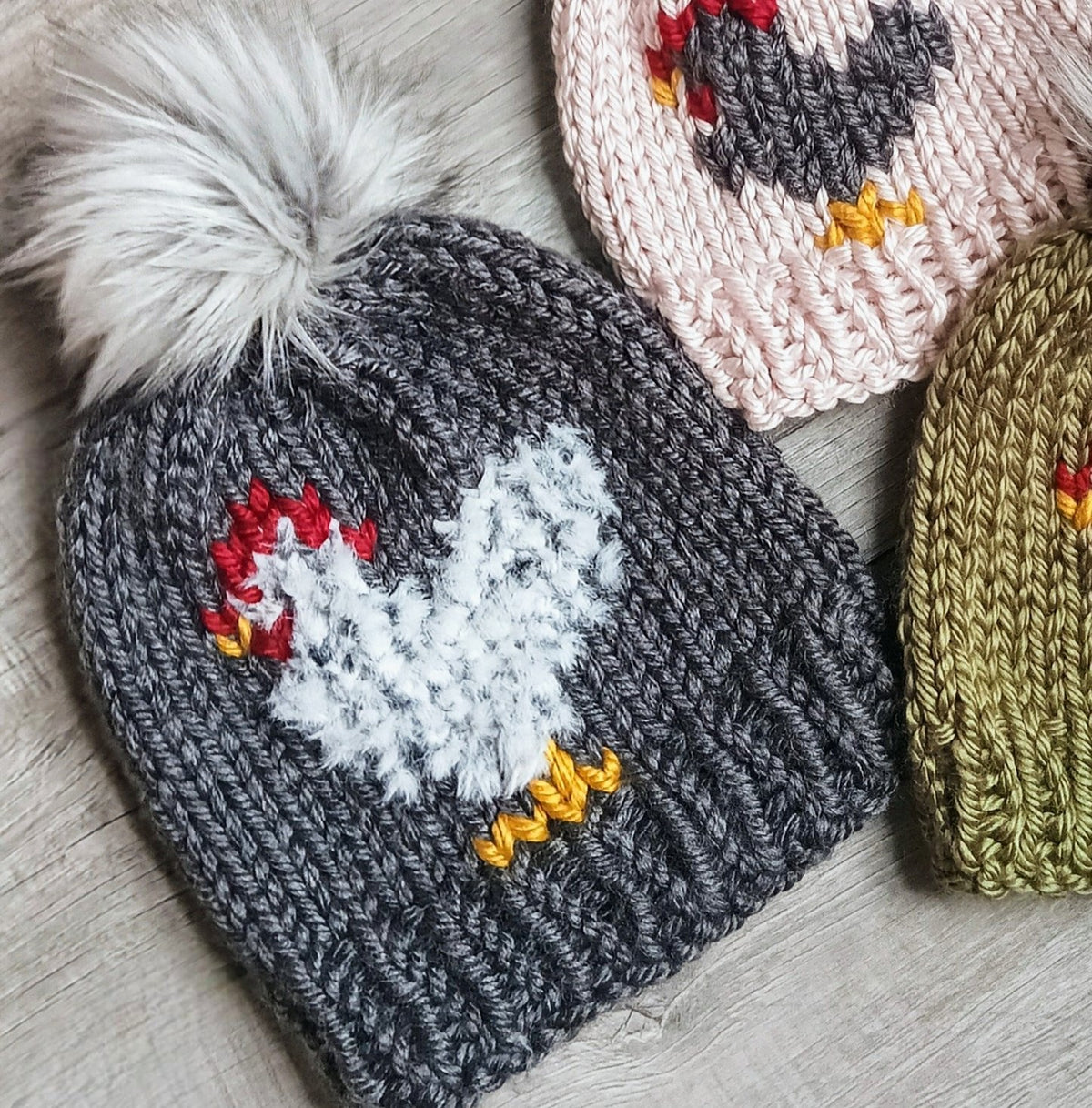 Charcoal knit beanie with white chicken embroidery, yellow accents, and faux fur pom-pom
