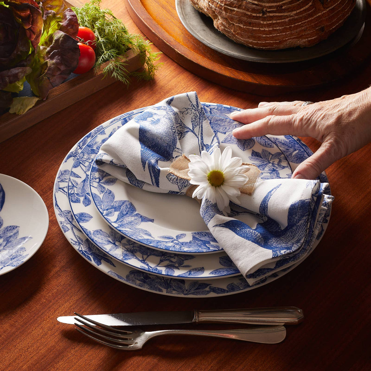Blue botanical porcelain dinner plate with white center, blue rim border; hand folds napkin
