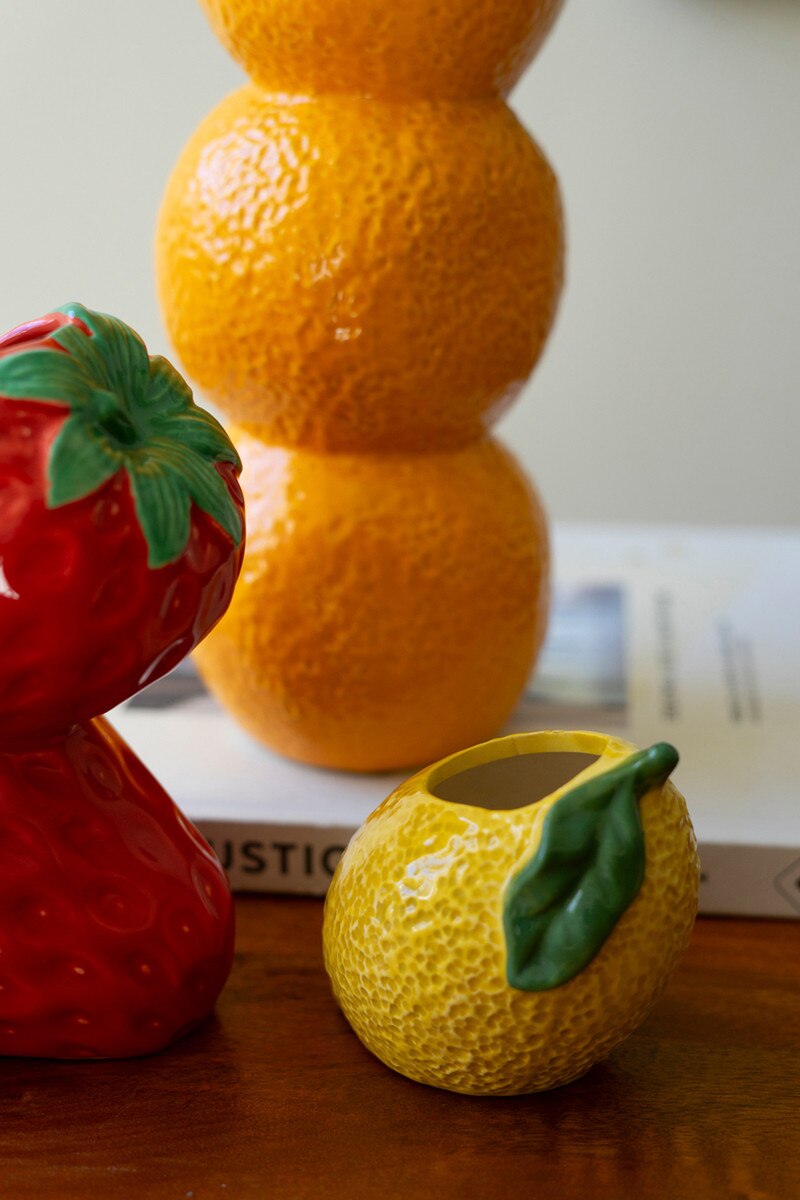 Ceramic fruit vases on table: red strawberry vase, small yellow bud vase with green leaf