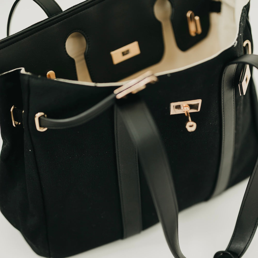 Black canvas tote bag with vegan leather trim, dual handles, gold hardware, front pockets.