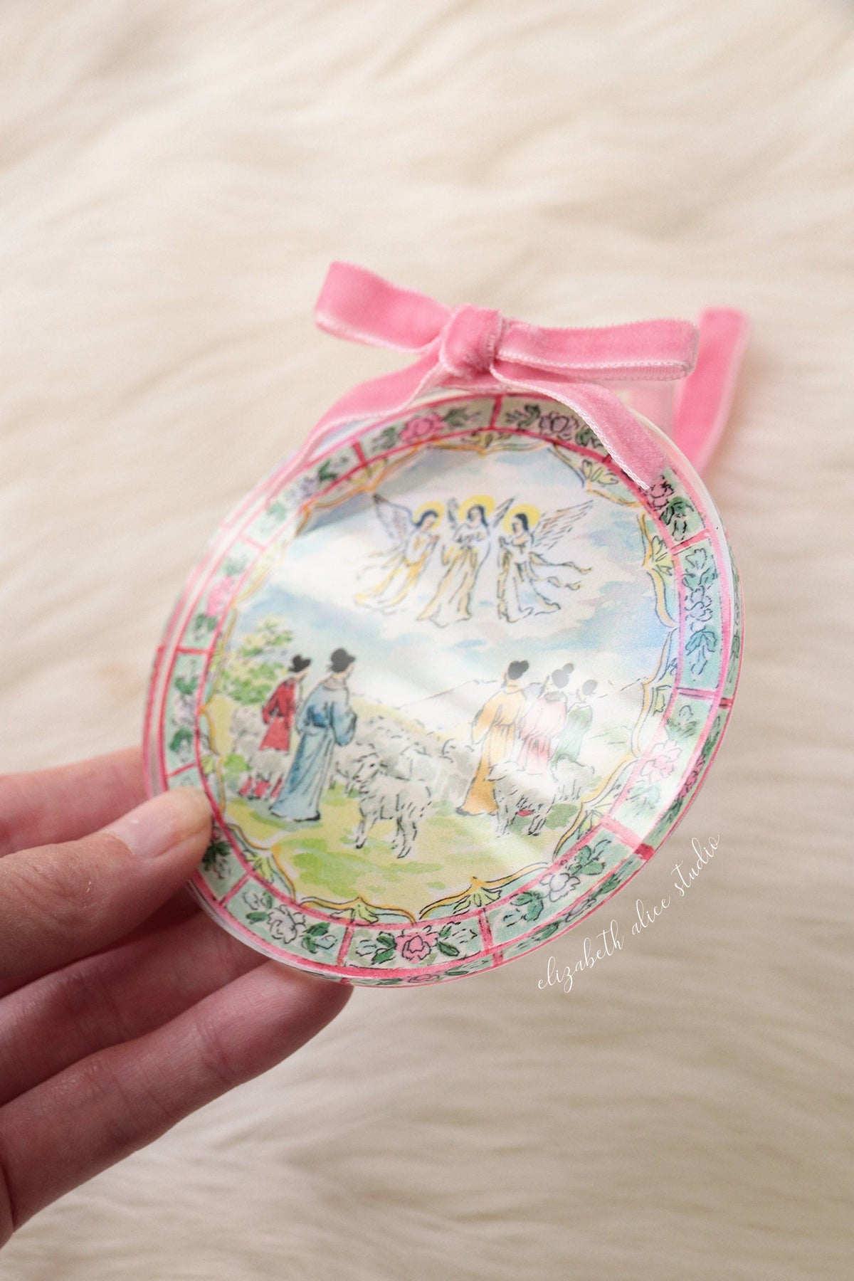 Round acrylic ornament depicting shepherds and angels, with a pink velvet bow and loop