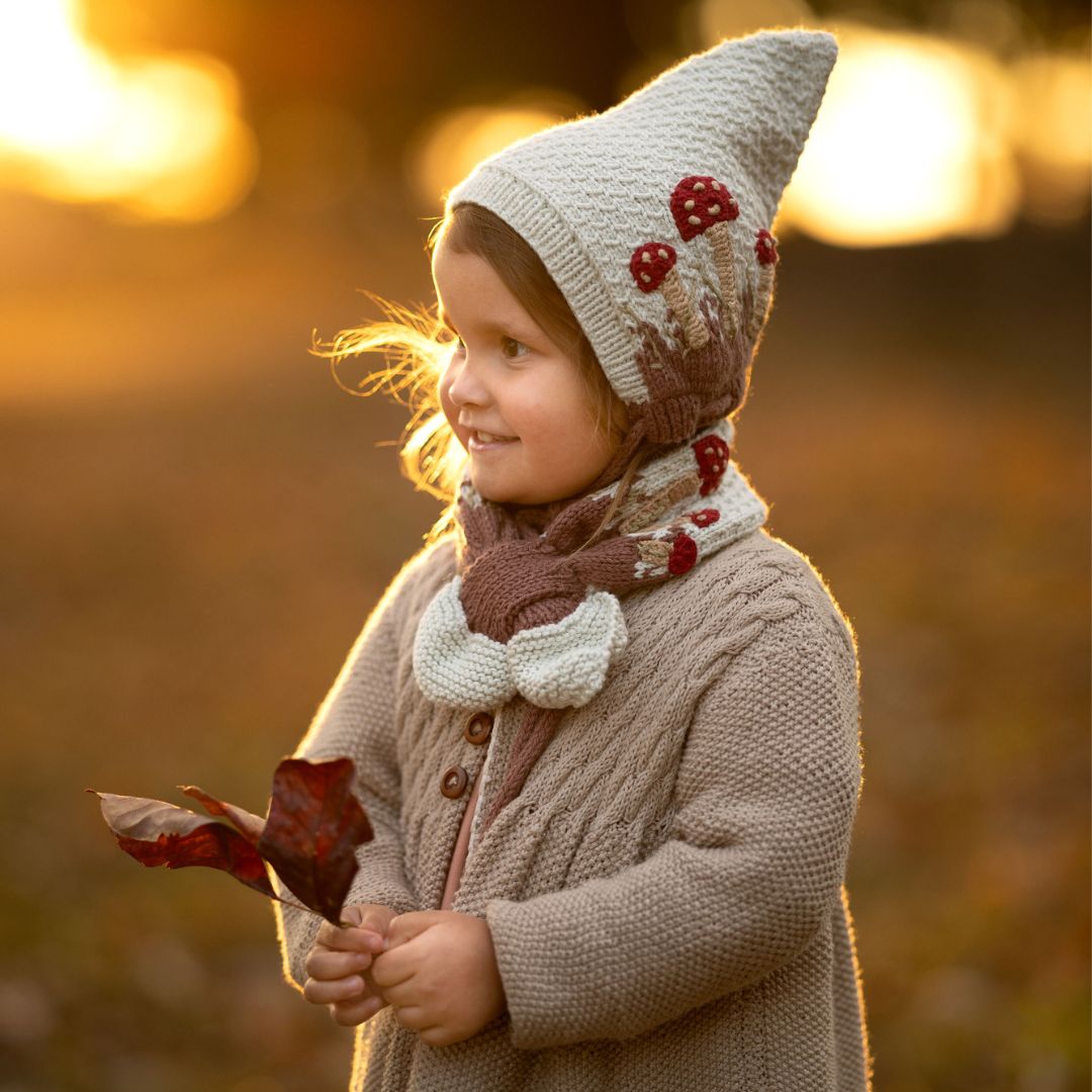 Cream knit pixie hat with red mushroom patches and brown tie detail