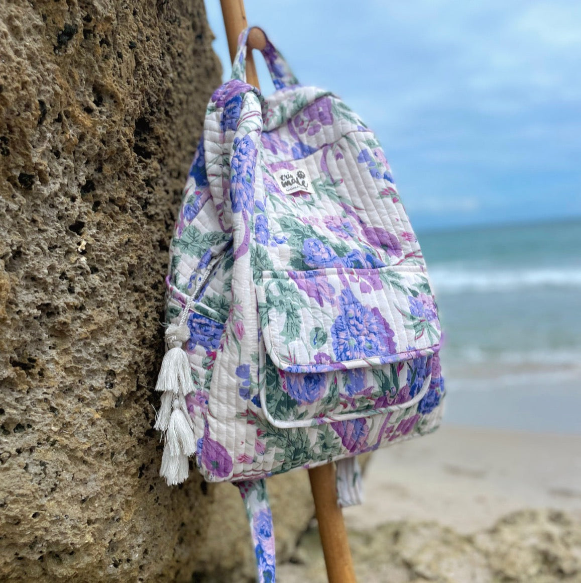 Quilted floral backpack with tassel on the left side, held near a rocky beach
