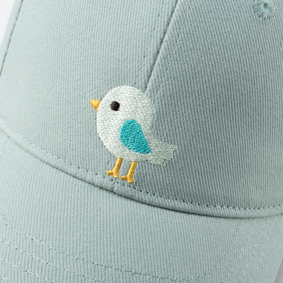 Close-up of the pale blue cap's embroidered white bird patch with teal wing and yellow feet