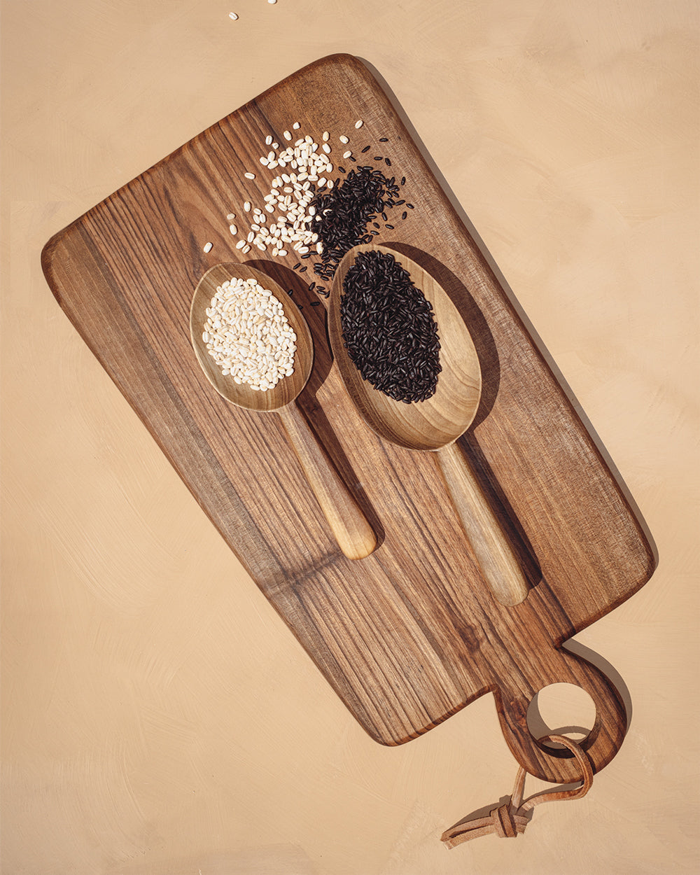 Two walnut scoop spoons on wood tray, filled with light and dark grains