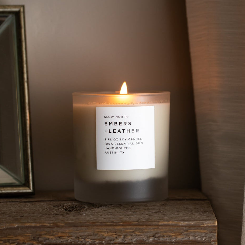 Embers + Leather scented candle in a frosted glass jar, lit with a white label.