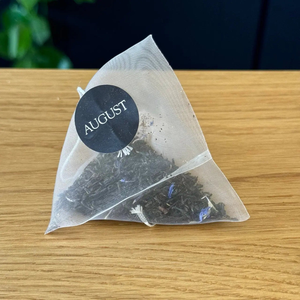 Triangular mesh Earl Grey tea bag with dark tea leaves and an AUGUST tag.