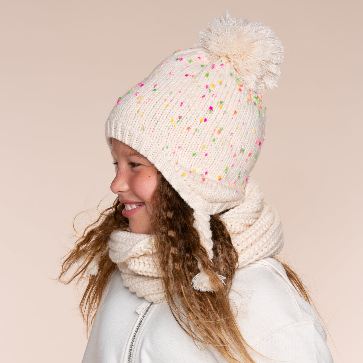 Side view of cream knit earflap hat with colorful flecks, pom-pom and braided tie