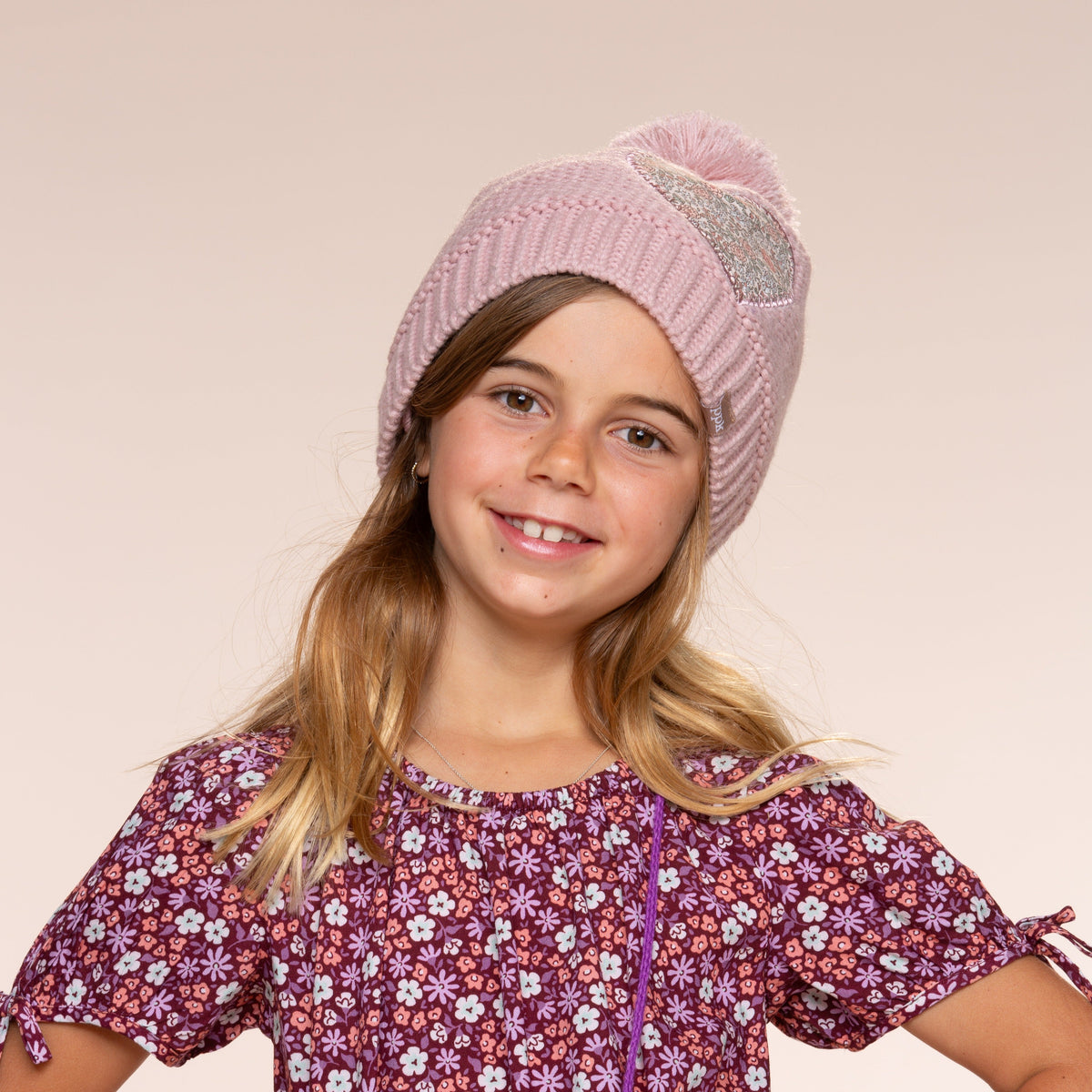 Pink knit beanie with pom-pom and floral heart patch, worn on a child