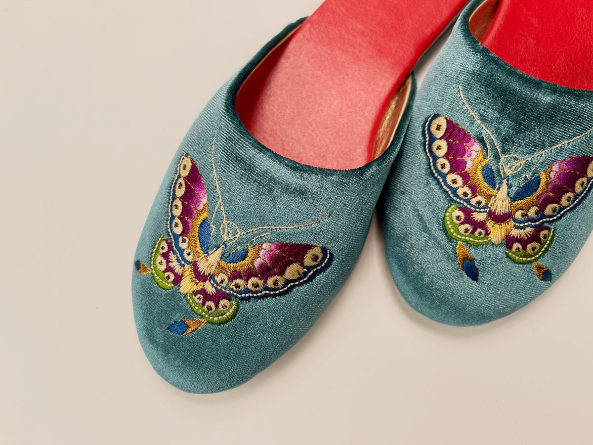 Close-up of teal velvet mule slippers with butterfly embroidery and red insoles