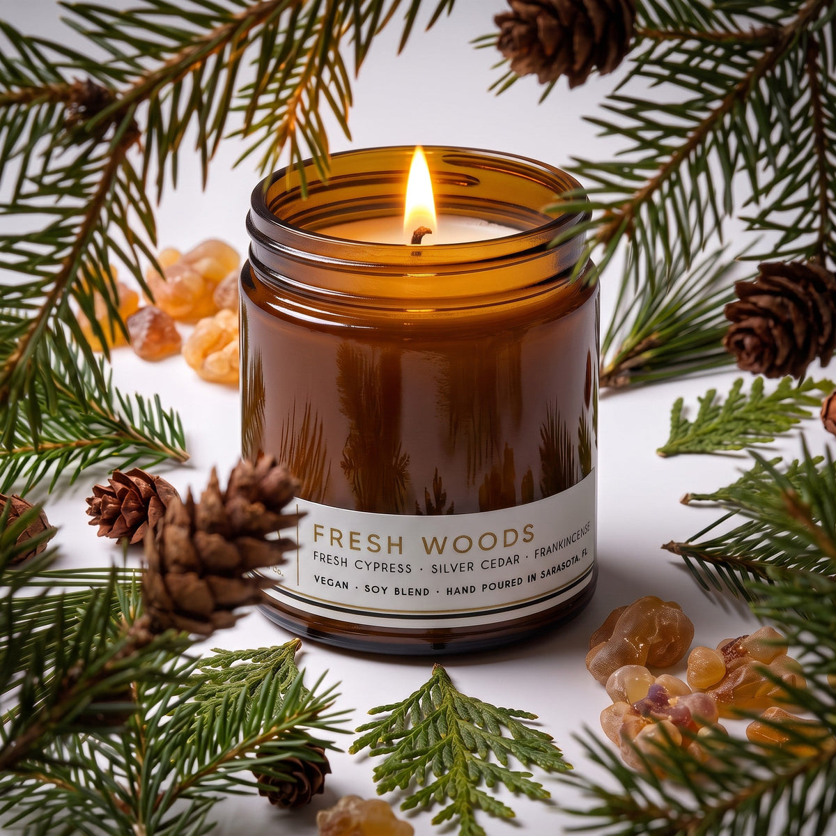 Lit Fresh Woods candle in an amber jar with a white label and evergreen sprigs