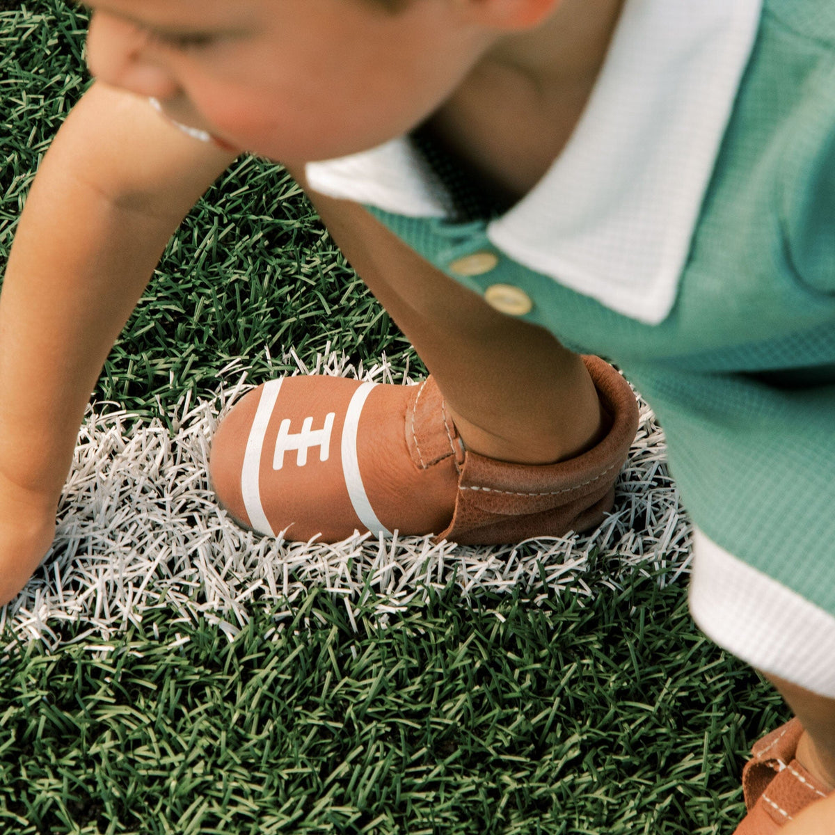 Brown baby shoes on child’s feet, hook-and-loop strap with H emblem