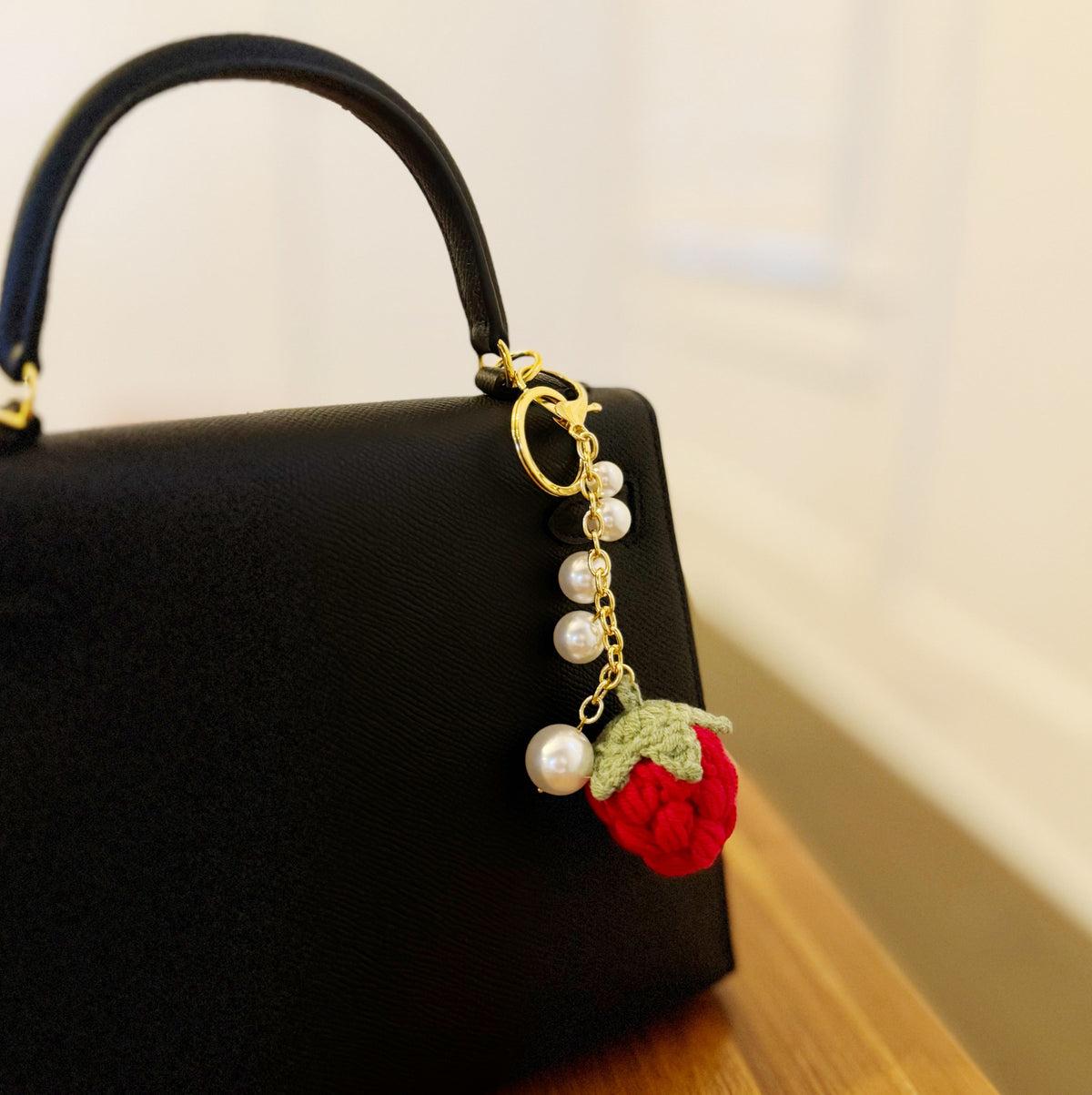Gold bag charm with red strawberry pom-pom and white pearls, shown on black bag handle