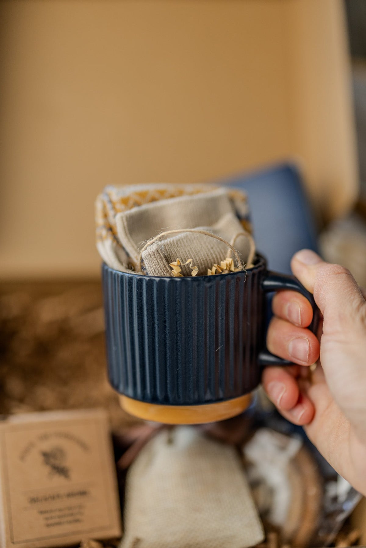 Hand holding navy ribbed mug from gift box; beige pouch and card below
