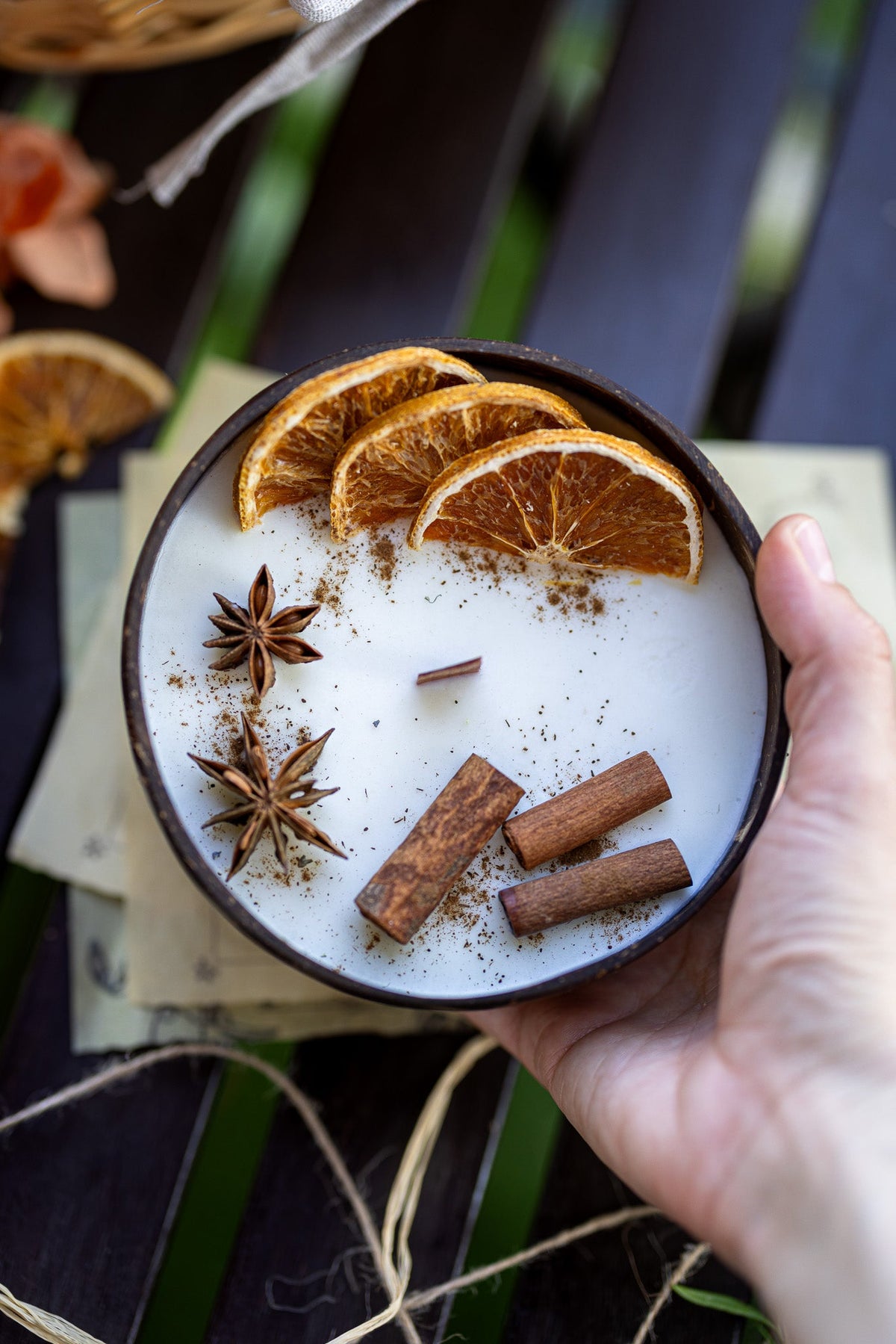 Coconut candle bowl held in hand with white wax, orange slices, cinnamon sticks, and star anise.