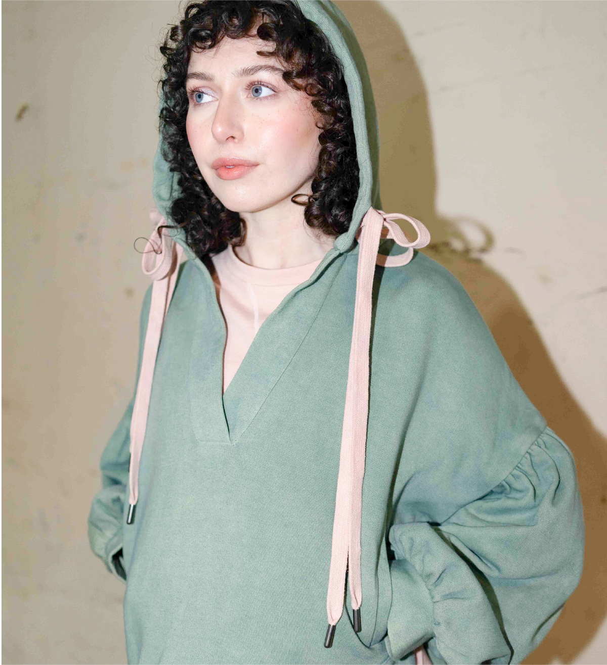 Sage green hoodie worn with the hood up, showing pink drawstrings and gathered sleeves.
