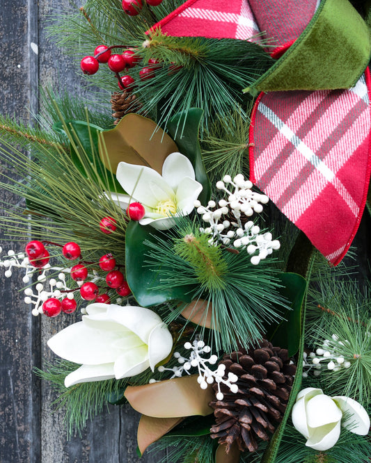 Close-up holiday wreath with red-and-white plaid bow, pine needles, pinecone, and white blooms