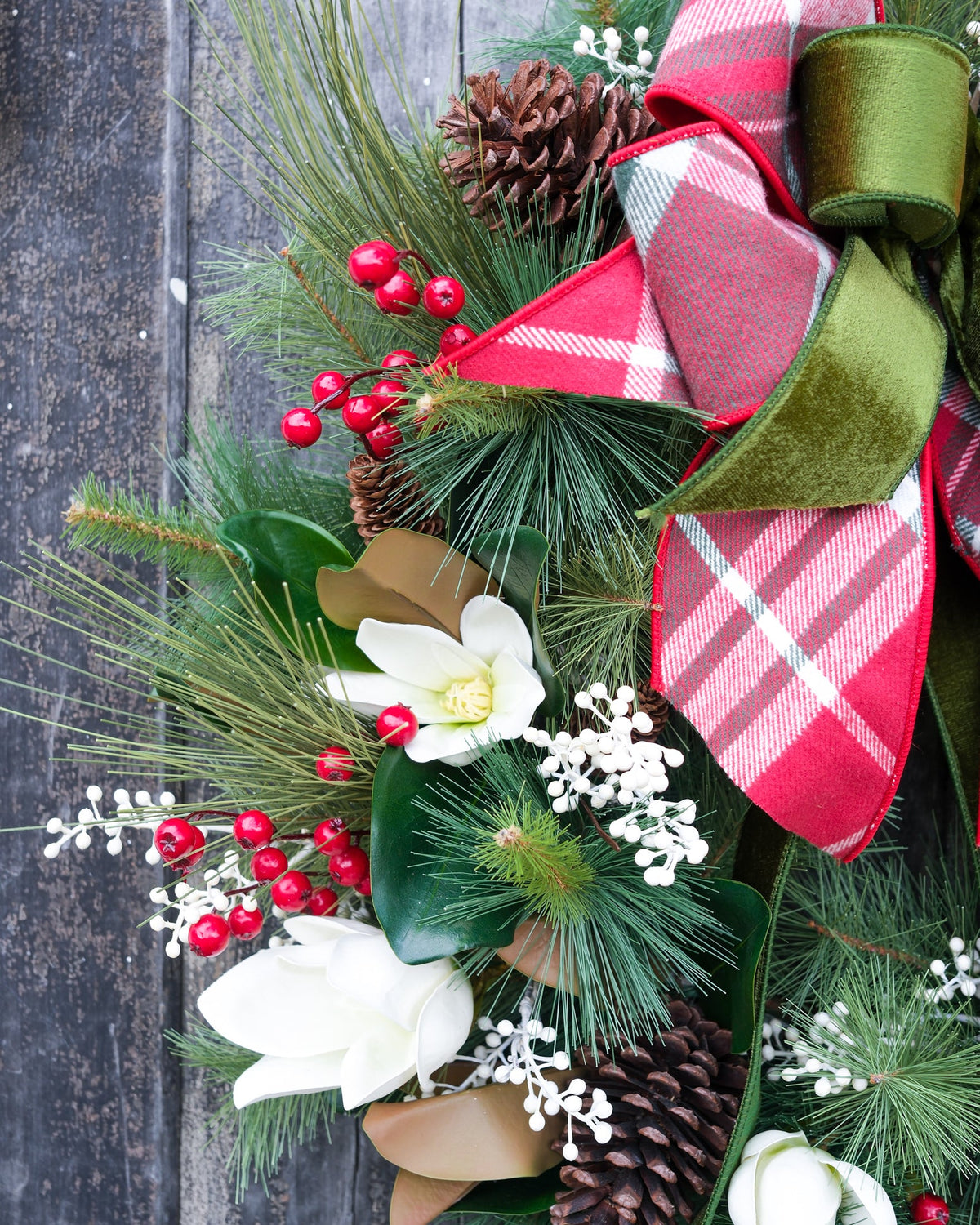 Holiday wreath close-up with red plaid bow, white blossoms, red berries, and pinecone