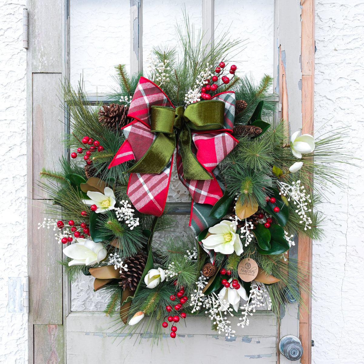 Door-hanging holiday wreath with green pine branches, white flowers, red berries, and plaid bow
