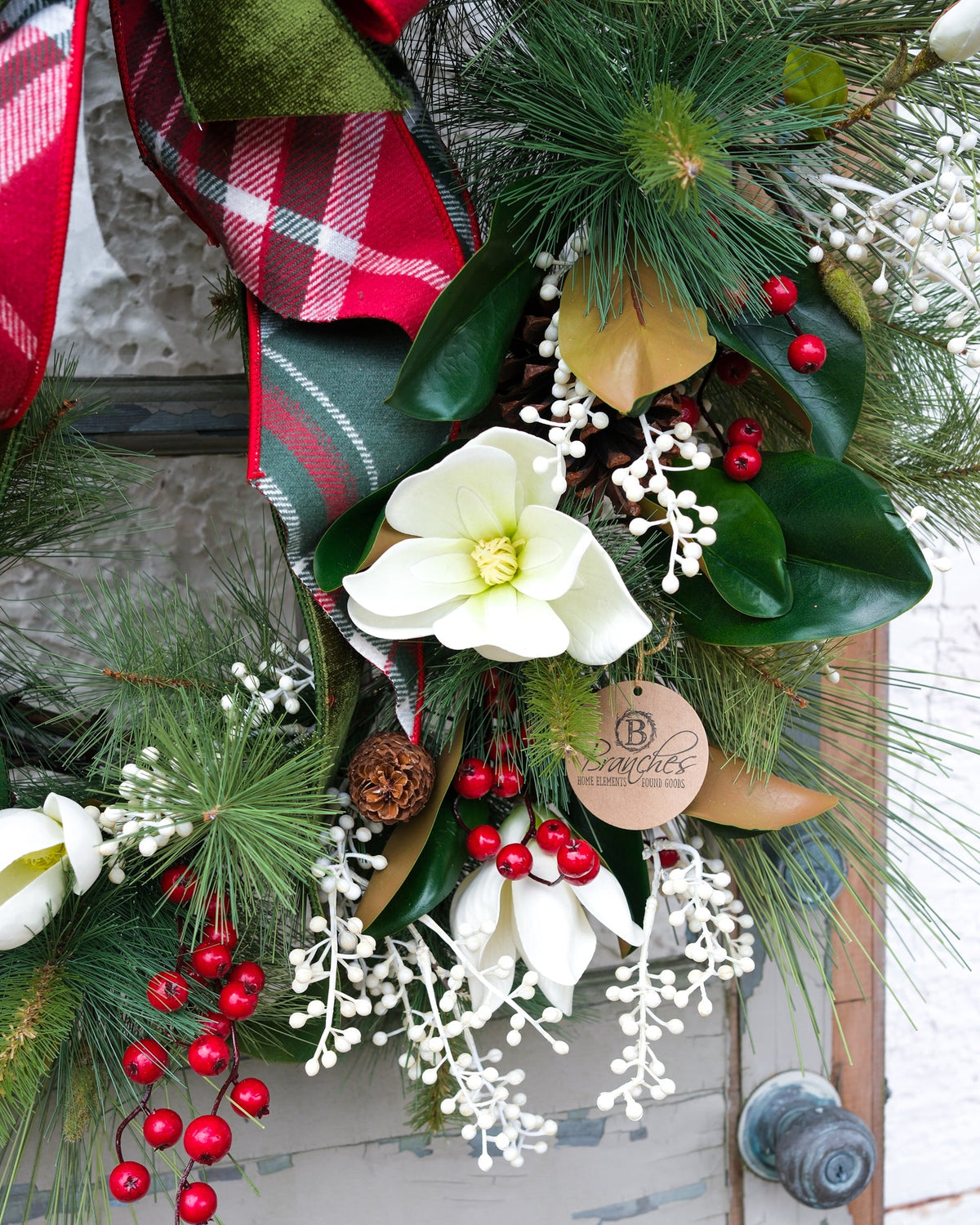 Holiday wreath detail with white flowers, pine needles, red berries, pinecone, and plaid ribbon