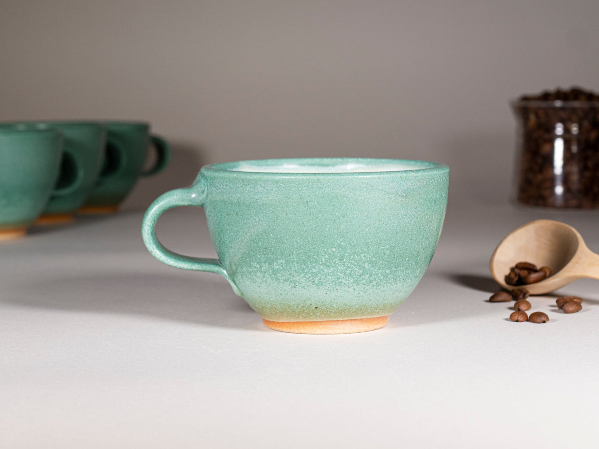 Green stoneware latte cup with handle, speckled glaze; set partially visible behind