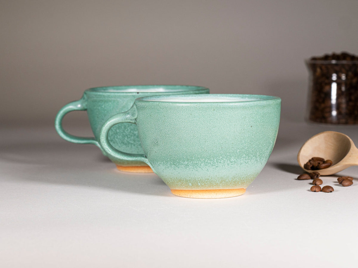 Green stoneware latte cup with handle, speckled glaze; saucer behind