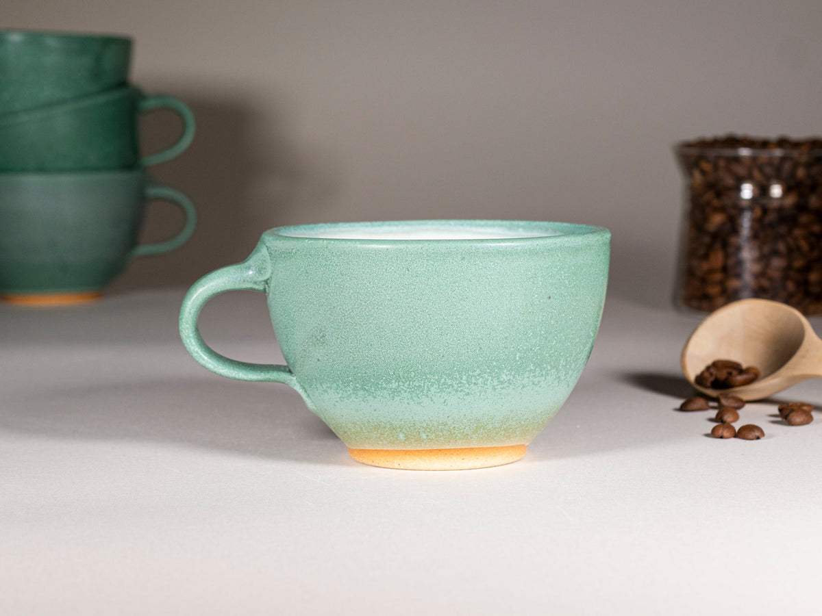 Green stoneware latte cup with handle, speckled glaze; partial set in background