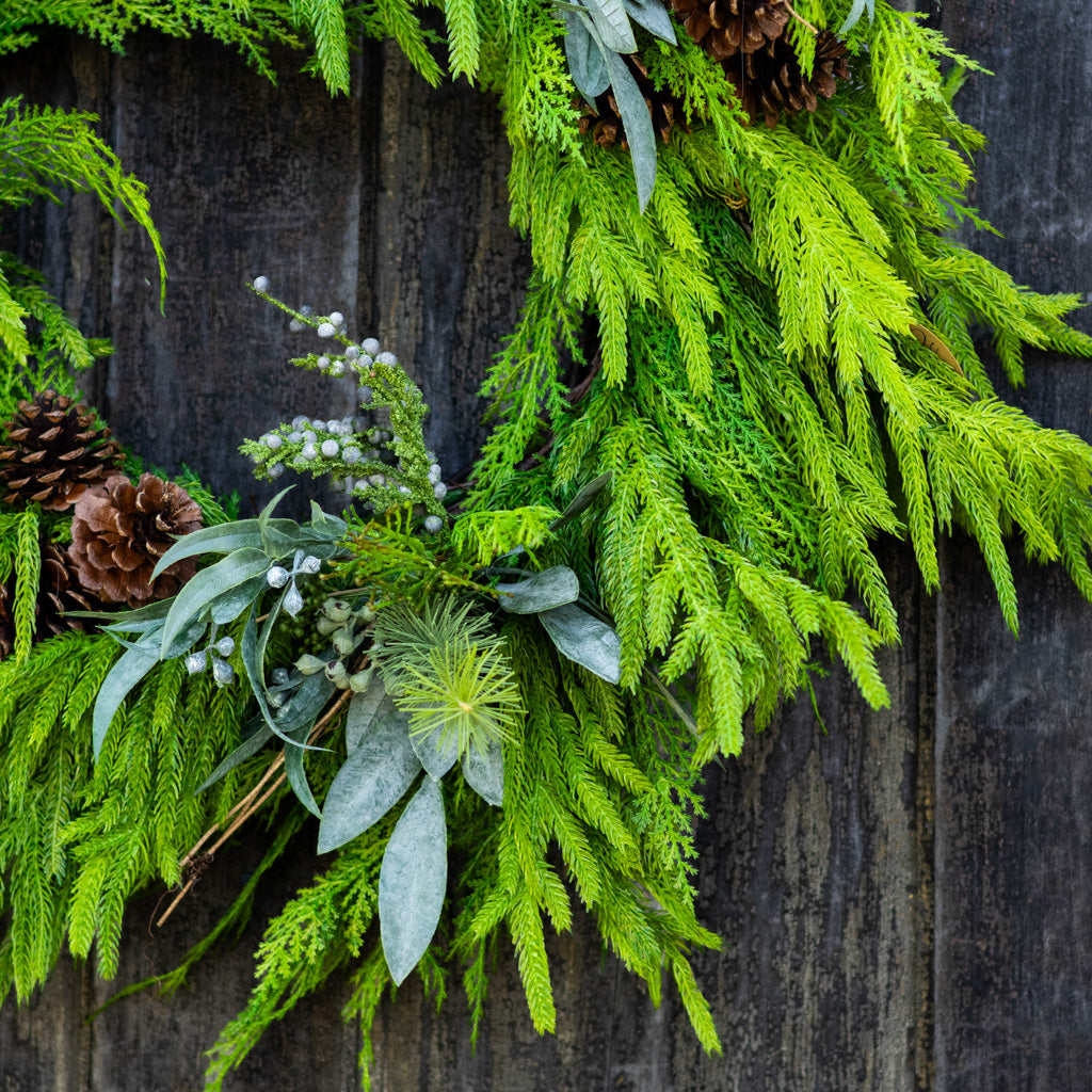 Green evergreen wreath detail with pale blue leaves, pinecone accents, and berry stems