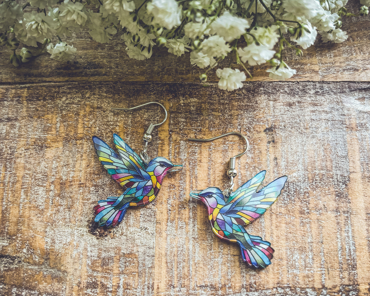 Colorful hummingbird dangle earrings with silver hooks on weathered wood surface.