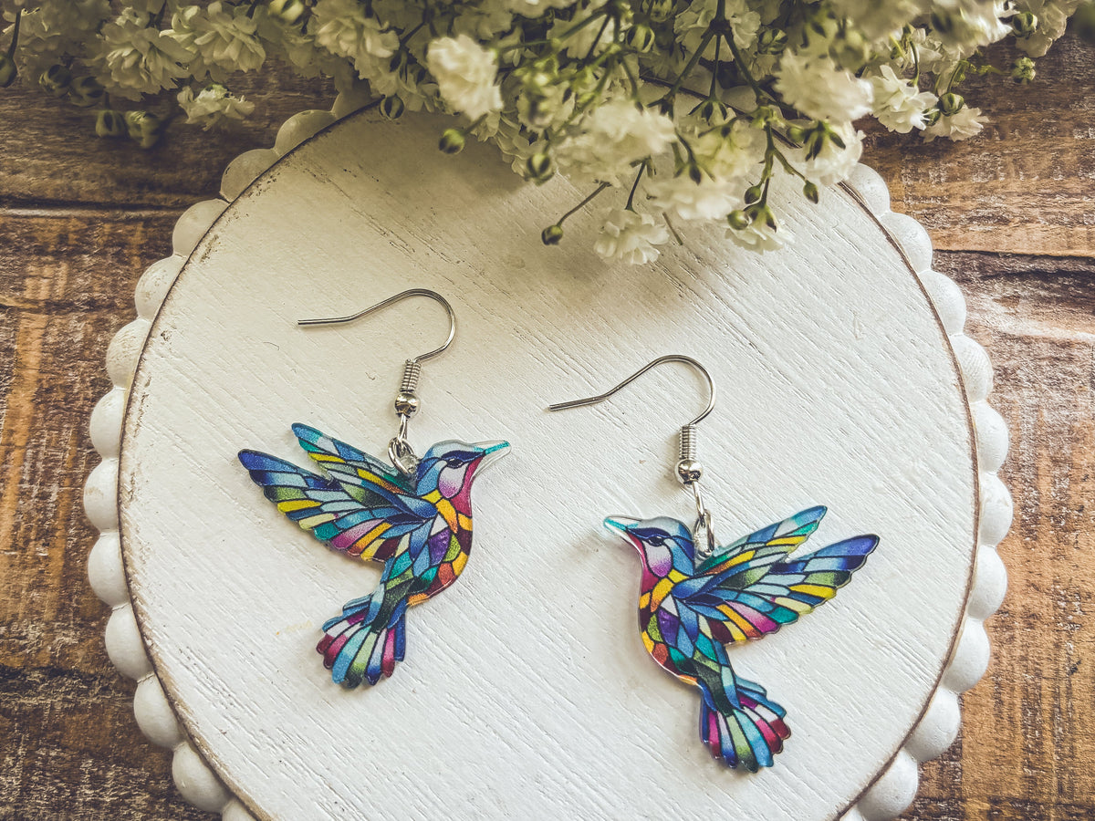 Colorful hummingbird dangle earrings with silver hooks on round white card.