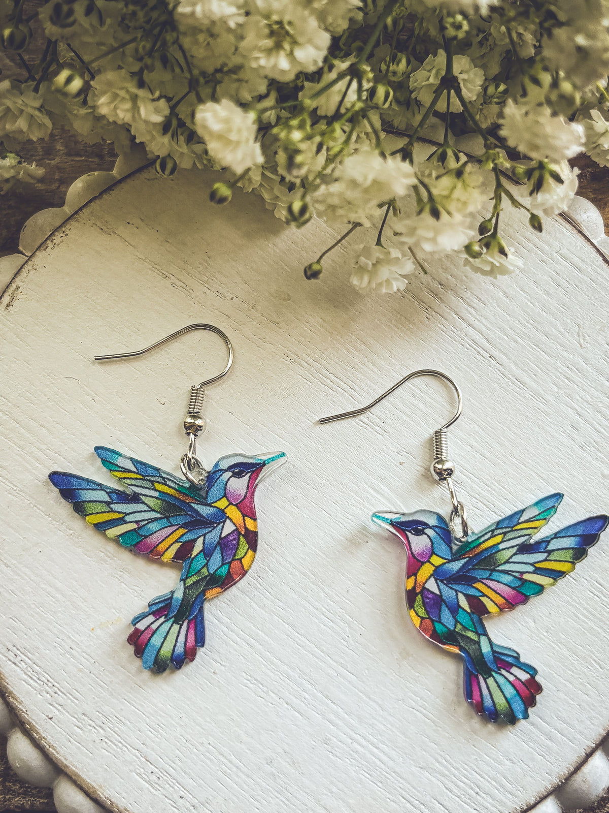 Colorful hummingbird dangle earrings with silver hooks on round display board.