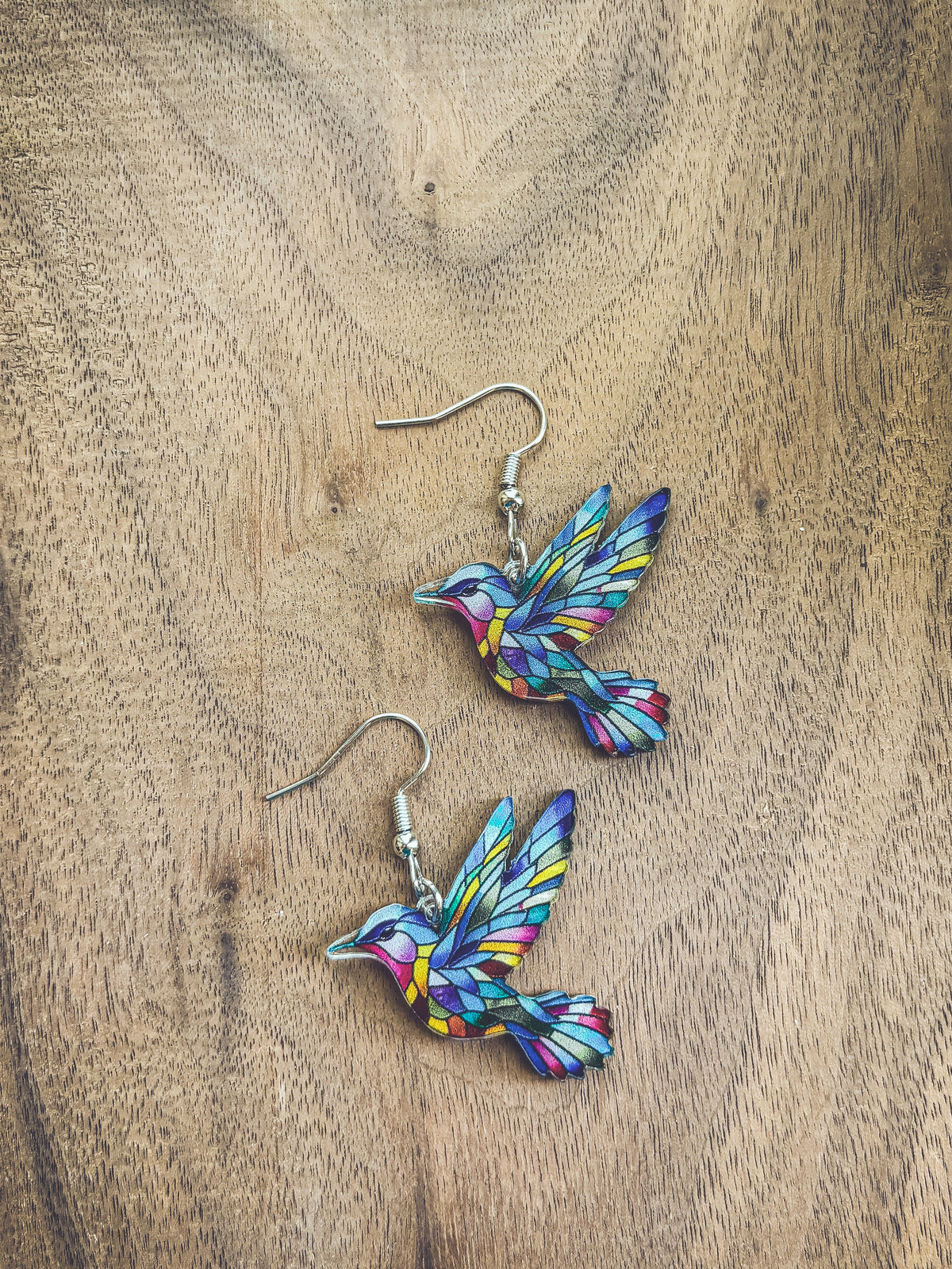 Colorful hummingbird dangle earrings with silver hooks on wood, two-piece view.