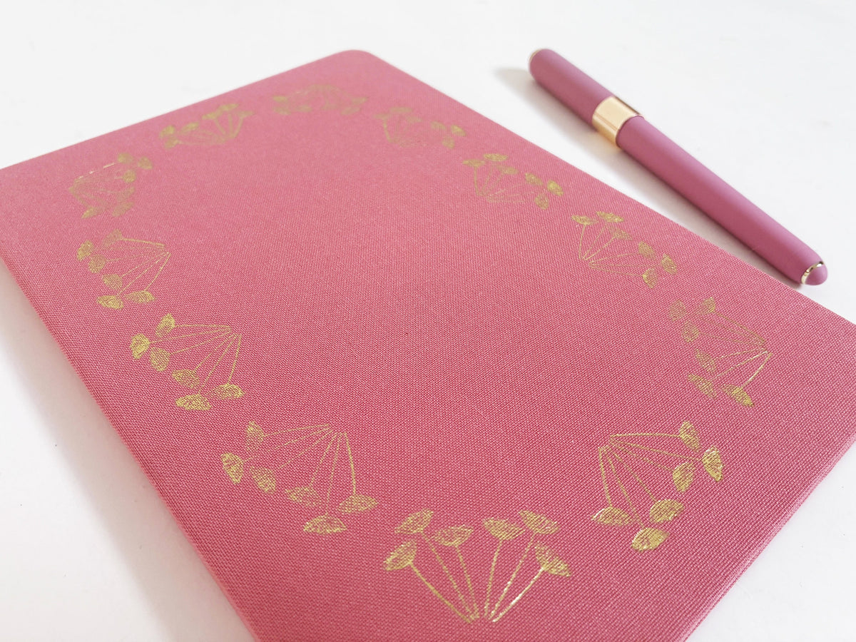 Pink linen-look notebook with gold foil floral border and ribbon; pen nearby