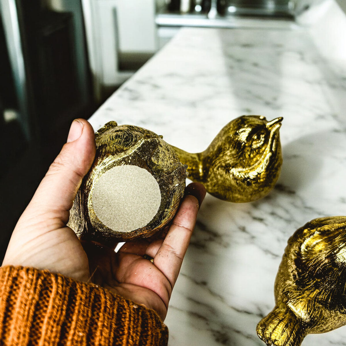 Hand holding a gold-tone bird figurine facing forward on a marble surface
