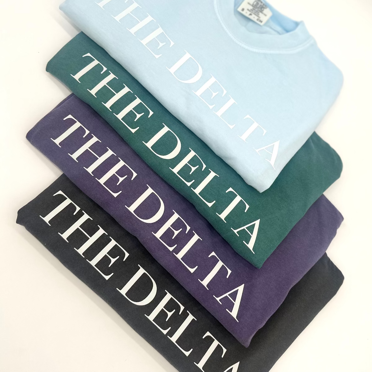 Folded graphic T-shirt set with “THE DELTA” lettering in light blue, purple, teal, and dark colors.