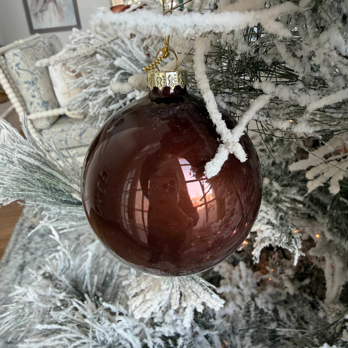 Round dark brown glass ornament with gold cap hanging from snowy branches