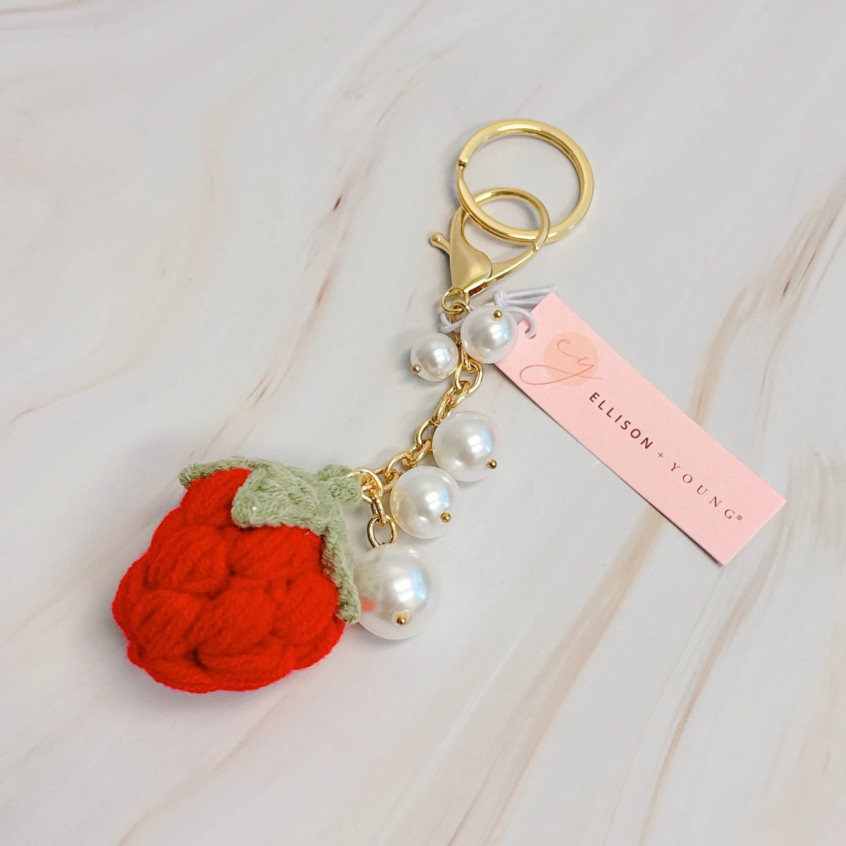 Gold bag charm with red strawberry pom-pom, green leaf, white pearls, and pink tag
