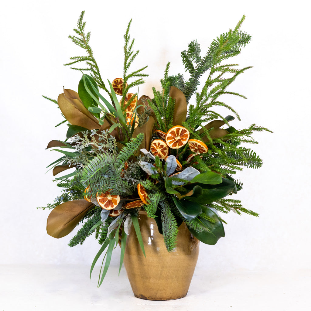 Holiday floral bouquet in a tan pot with evergreen sprigs, ribbon, and orange slice decorations.