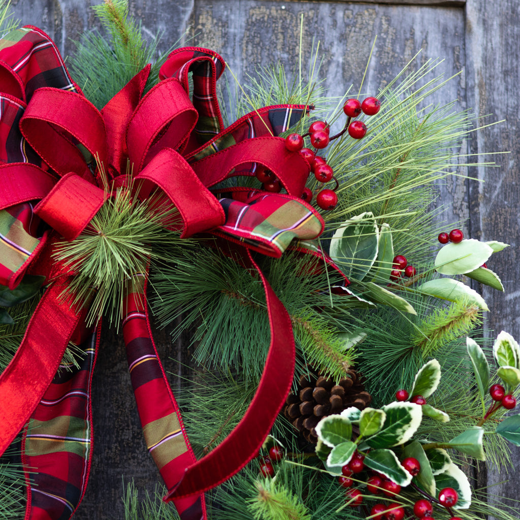 Holiday wreath corner view with red ribbon loops, evergreen branches, berries, and pinecones
