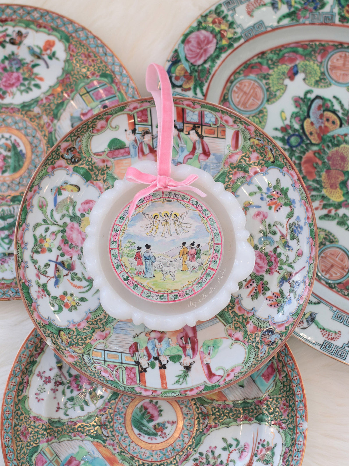 Scalloped acrylic ornament with shepherds and angels scene, pink ribbon, shown against decorative plates