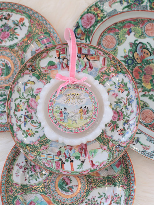 Scalloped acrylic ornament with shepherds and angels scene, pink ribbon, shown against decorative plates