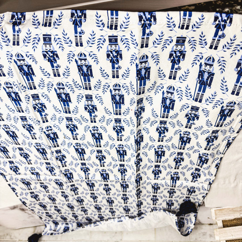 White tea towel covered in blue nutcracker and leaf print with navy tassel corners