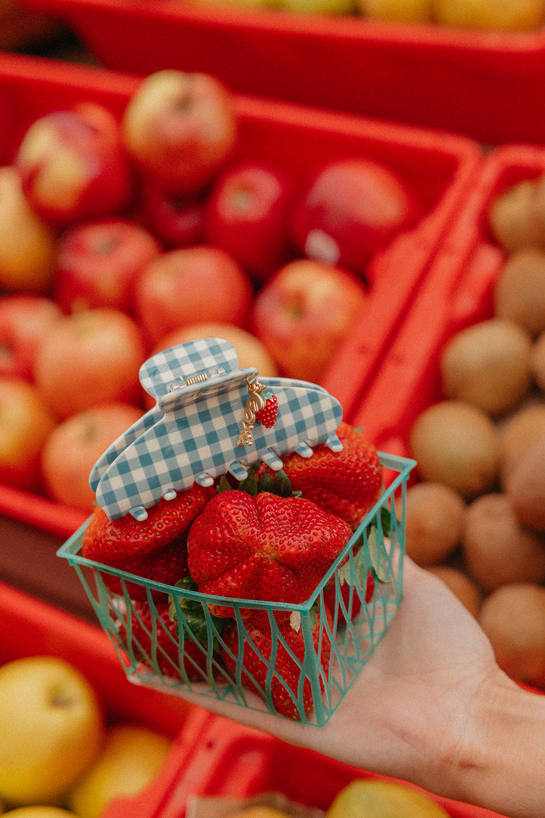 Blue gingham claw hair clip with red strawberry charm resting in fruit display.