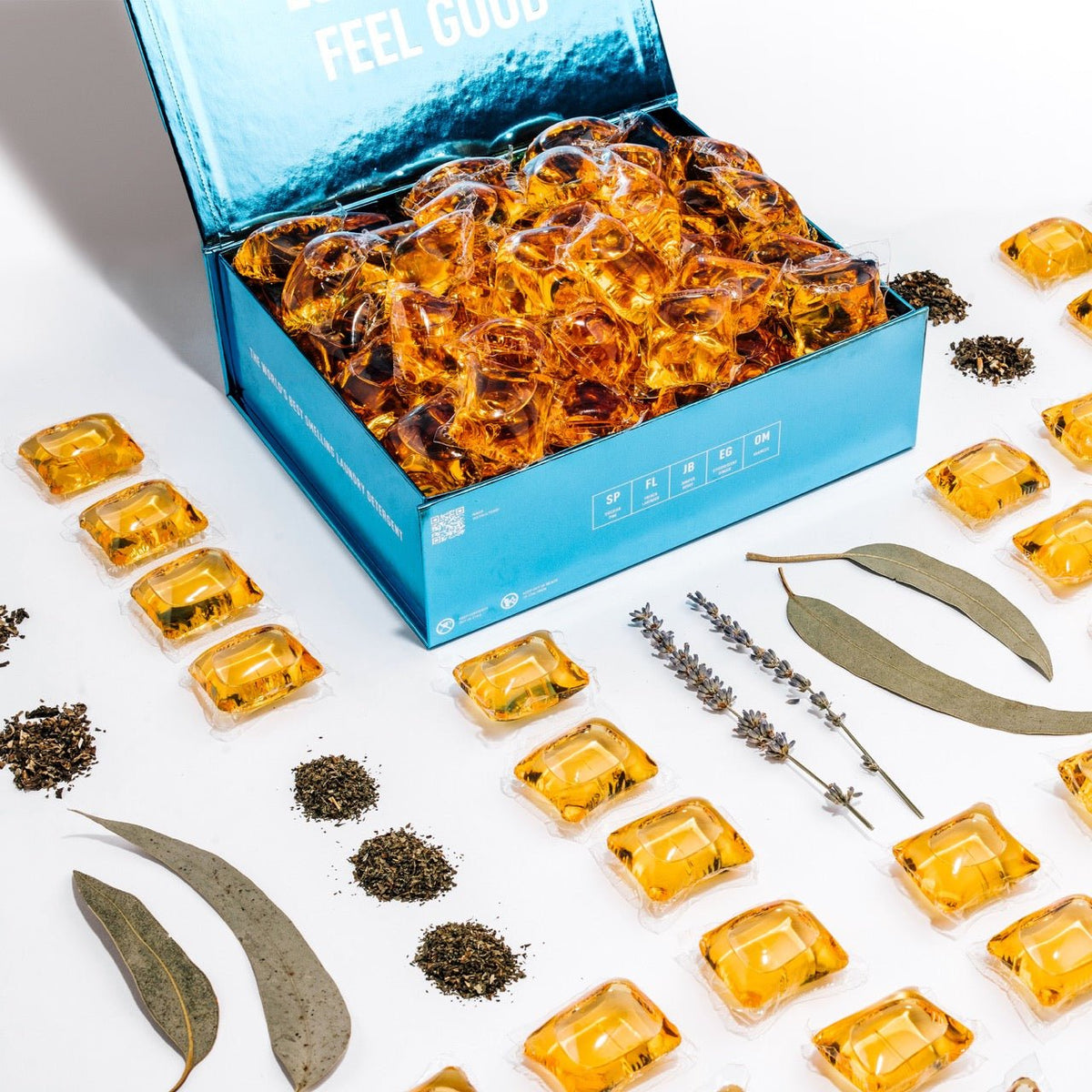 Blue open box of laundry detergent pods with amber pods and dried herb pieces