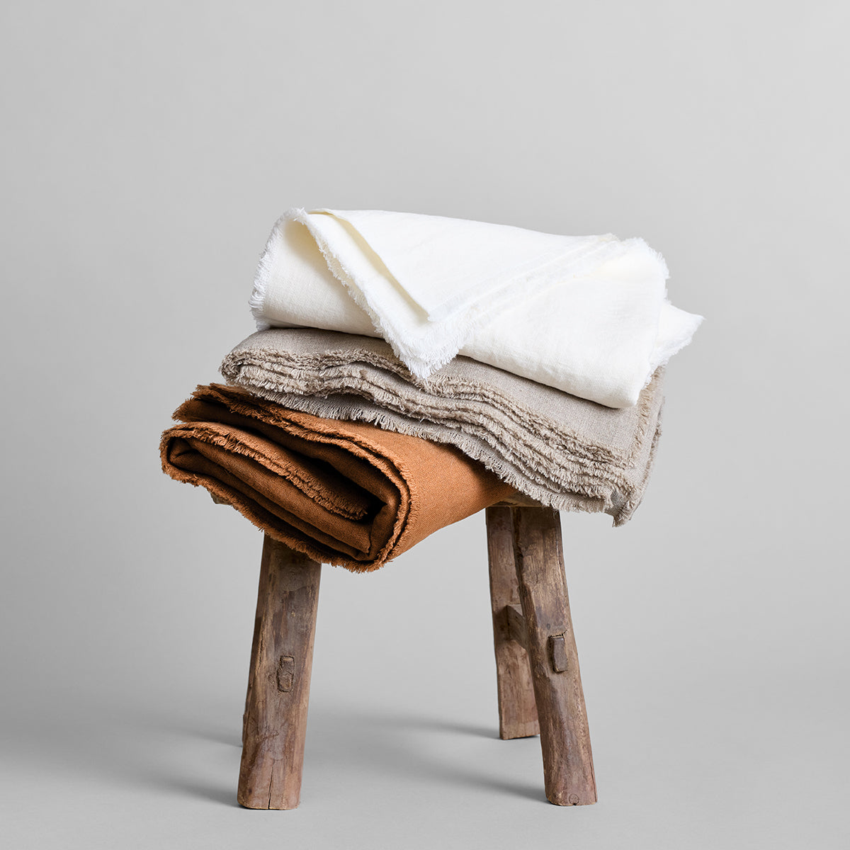 Fringed linen tablecloth stack in white, gray, and tan on a wooden stool