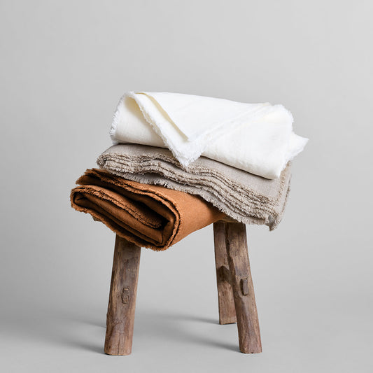 Fringed linen tablecloth stack in white, gray, and tan on a wooden stool