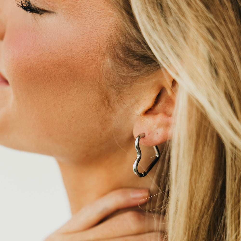 Silver-tone heart hoop earrings, close-up on an earlobe with open heart design.