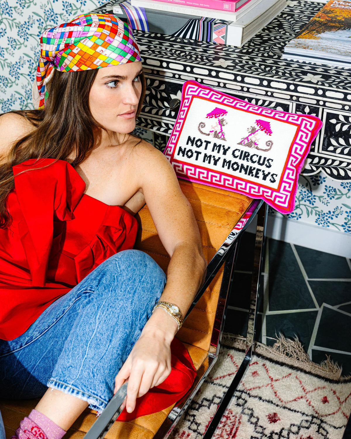 Needlepoint throw pillow shown beside a seated woman, with Greek key border and slogan